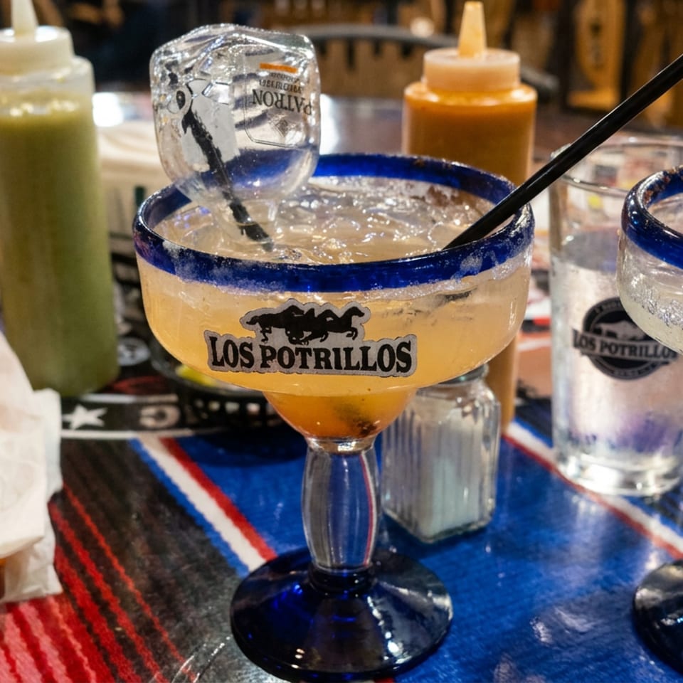 Two Los Potrillos margaritas with blue rims sit on a colorful restaurant table beside sauces, chips, napkins, and utensils.
