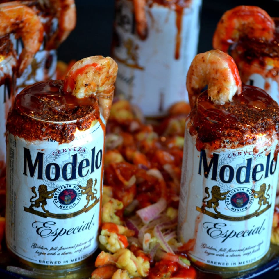 Modelo Especial beer cans rimmed with chili and sauce, topped with shrimp and surrounded by a colorful seafood dish.