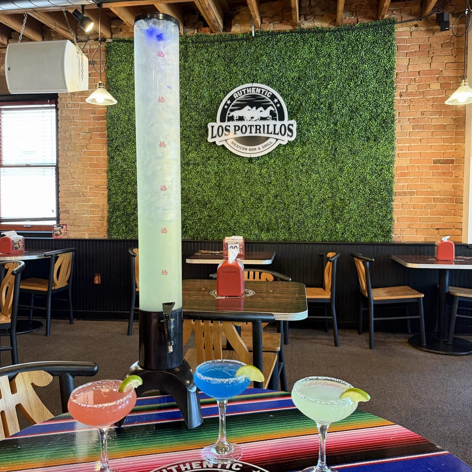 Three colorful margaritas sit on a striped table at Los Potrillos Mexican Bar & Grill, with rustic brick walls and restaurant seating behind them.