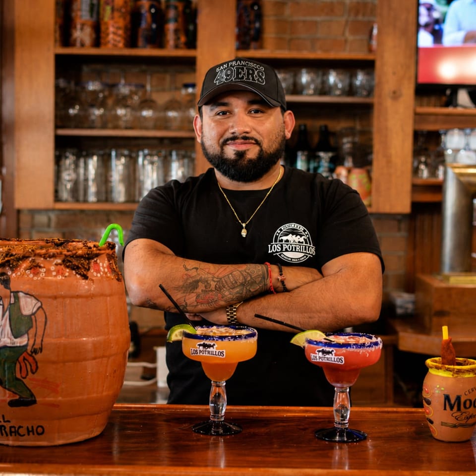 Bartender in a Los Potrillos shirt poses behind a bar with colorful margaritas and a large decorated clay drink vessel.