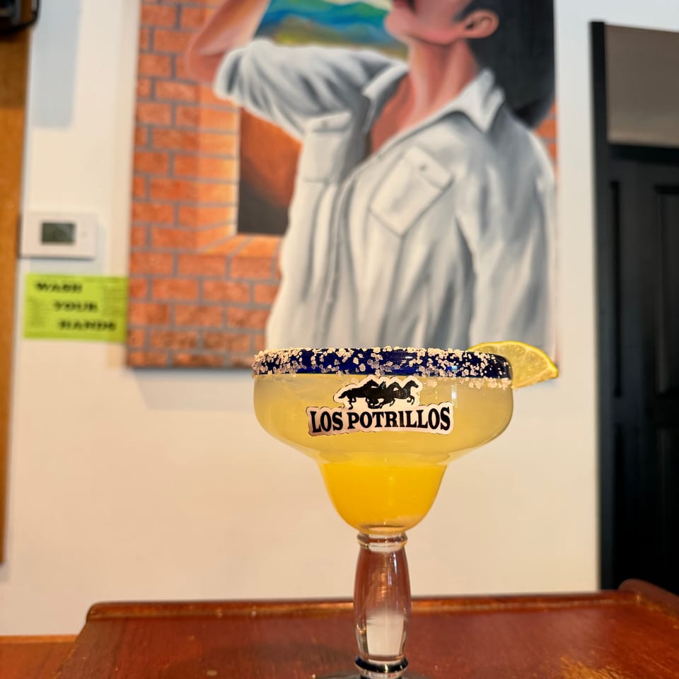 Salt-rimmed margarita with a lime wedge and Los Potrillos branding sits on a wooden table before a colorful mural.