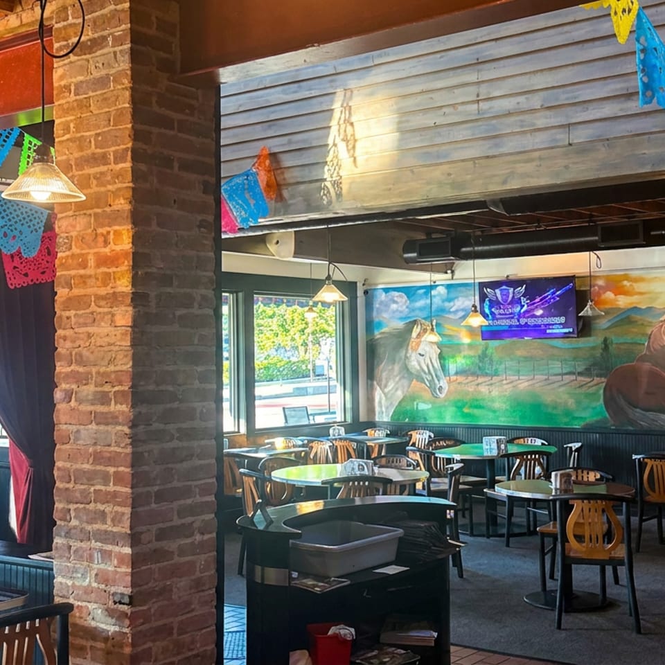 Colorful restaurant dining room with papel picado banners, horse mural, exposed brick, pendant lights, tables, and a seated woman.