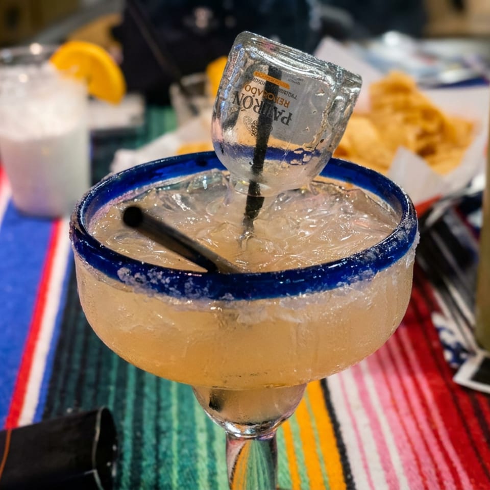 Blue-rimmed margarita glass with ice and upside-down miniature bottle on a colorful striped restaurant table.