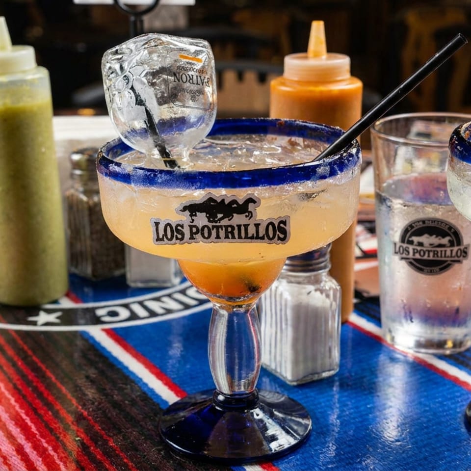 Los Potrillos restaurant table with two blue-rimmed margaritas, a branded tumbler, salsa bottles, chips, and colorful woven textiles.