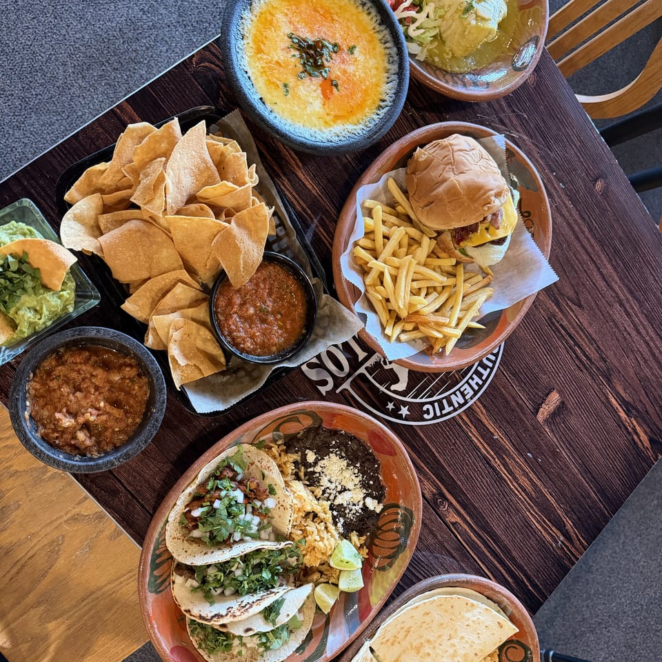 Overhead view of a Mexican restaurant spread with tacos, quesadillas, rice, beans, burger, fries, chips, salsa, guacamole, and queso dip.