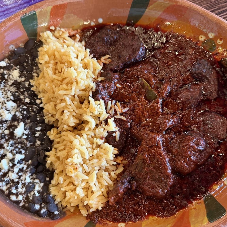 Mexican-style plate with black beans topped with queso fresco, yellow rice, and saucy grilled steak in a colorful ceramic dish.