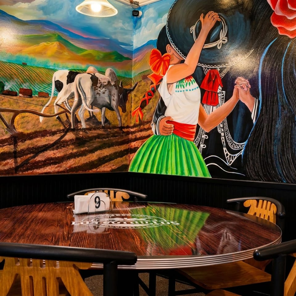 Restaurant dining table beneath a colorful mural depicting Mexican dancers, farmland, cattle, and a decorated skull.
