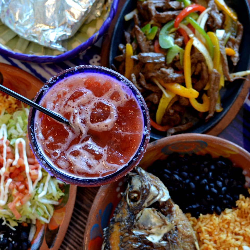 Overhead spread of Mexican dishes with rice, beans, salad, grilled peppers and beef, centered around an iced red drink.